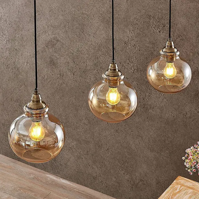 Lindby Pendelleuchte Tymoni - creme / amber, amber, altmessing, E27 - Drei bernsteinfarbene Pendelleuchten aus Glas mit Messingbeschlägen hängen an einer Wand und beleuchten einen Raum.