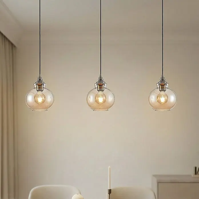 Lindby Pendelleuchte Tymoni - creme / amber, amber, altmessing, E27 - Drei Pendelleuchten mit bernsteinfarbenen Glaskugeln und sichtbaren Filamentbirnen beleuchten einen Essbereich.