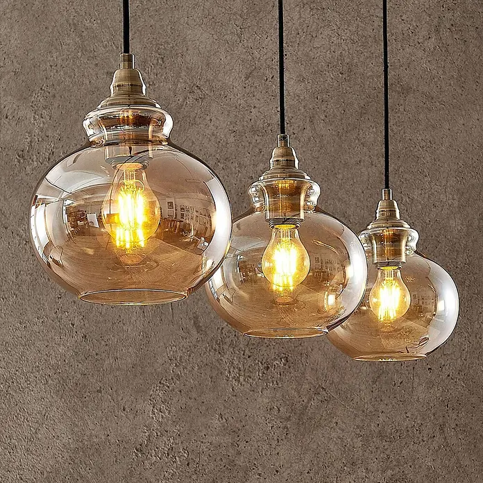 Lindby Pendelleuchte Tymoni - creme / amber, amber, altmessing, E27 - Drei bernsteinfarbene Glaspendelleuchten mit Messingbeschlägen und LED-Glühbirnen an einer strukturierten Wand.
