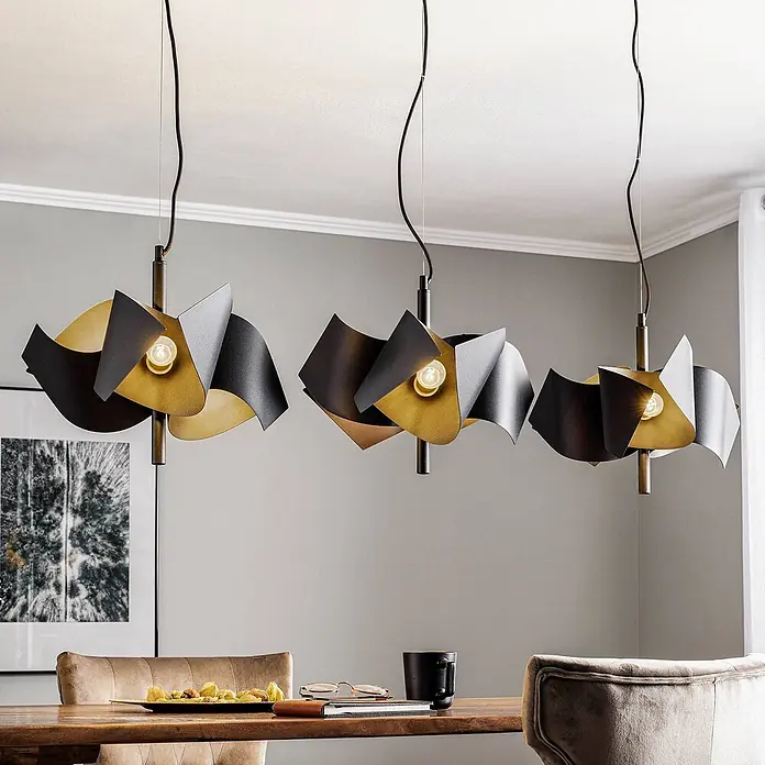 Lucande Pendelleuchte Imron - L x B x H: 142 x 40 x 35 cm, schwarz, sandSchwarz, E14 - Drei moderne Pendelleuchten mit geschwungenen schwarzen, goldenen und silbernen Metallplatten hängen über einem Holzesstisch.