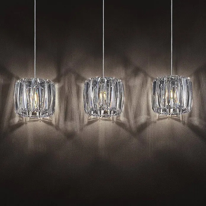 Lindby Pendelleuchte Sofia - L x B x H: 107 x 23 x 20 cm, klar / transparent, transparent, chrom, E14 - Drei beleuchtete Kristall-Pendelleuchten hängen vor einer dunklen, strukturierten Wand und werfen Lichtmuster.