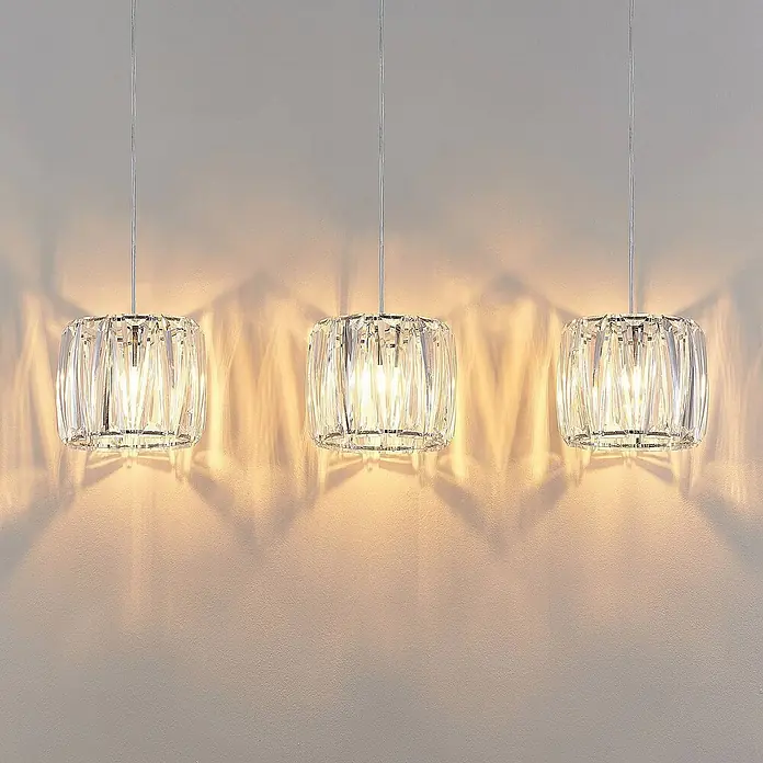 Lindby Pendelleuchte Sofia - L x B x H: 107 x 23 x 20 cm, klar / transparent, transparent, chrom, E14 - Drei moderne Pendelleuchten aus klarem, facettiertem Kristall hängen von der Decke und erzeugen warme Lichtmuster an der Wand.