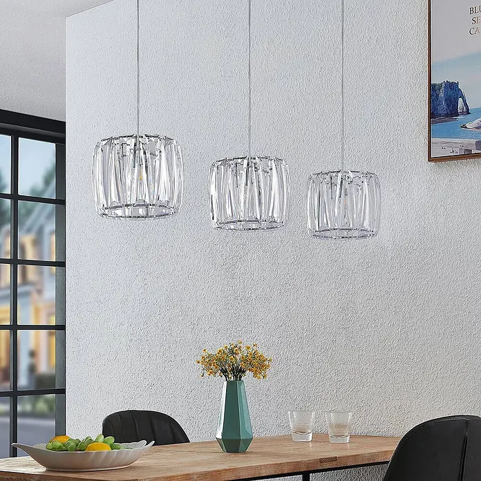Lindby Pendelleuchte Sofia - L x B x H: 107 x 23 x 20 cm, klar / transparent, transparent, chrom, E14 - Drei klare Kristall-Pendelleuchten hängen über einem Esstisch aus Holz und beleuchten den Raum.