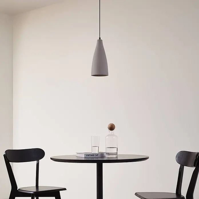 Lindby Pendelleuchte Sanne - alu / grau / zink, betongrau, E27 - Graue Beton-Pendelleuchte hängt über einem modernen Esstisch mit Stühlen, Buch und Glaswaren.
