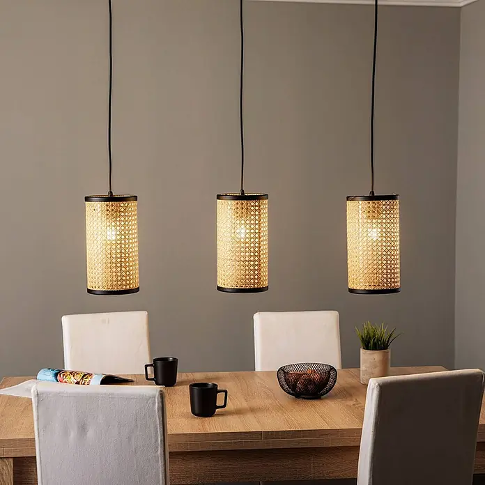 Lindby Pendelleuchte Yaelle - L x B x H: 92 x 15 x 28 cm, Holz hell, holz hell, E27 - Drei zylindrische Pendelleuchten aus Rattan und schwarzem Metall hängen über einem Holzesstisch.