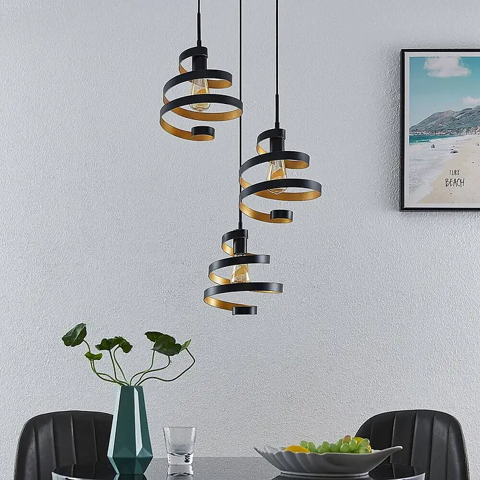 Lindby Pendelleuchte Colten - schwarz, mattSchwarz, gold, E27 - Drei schwarz-goldene Spiral-Pendelleuchten hängen in verschiedenen Höhen über einem Esstisch und zeigen modernes Design.