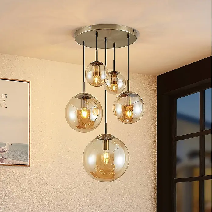 Lindby Pendelleuchte Teeja - L x B x H: 50 x 50 x 30 cm, creme / amber, amber, altmessing, E27 - Fünfflammige Pendelleuchte mit bernsteinfarbenen Glaskugeln unterschiedlicher Größe, von einer gebürsteten Metall-Deckenplatte in einem Raum hängend.