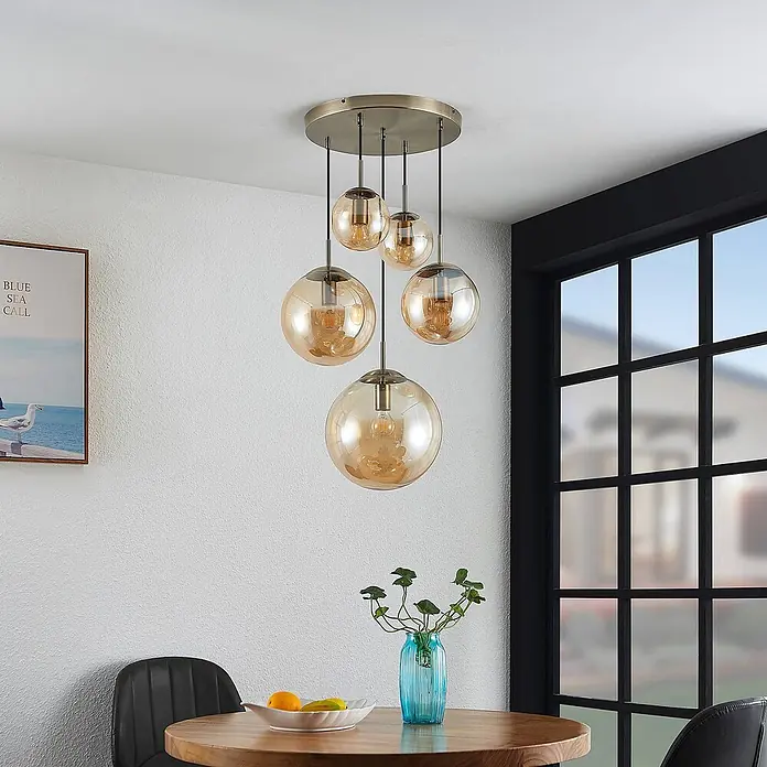 Lindby Pendelleuchte Teeja - L x B x H: 50 x 50 x 30 cm, creme / amber, amber, altmessing, E27 - Fünf bernsteinfarbene Glaskugelleuchten hängen von einer Deckenplatte aus gebürstetem Messing über einem Esstisch.