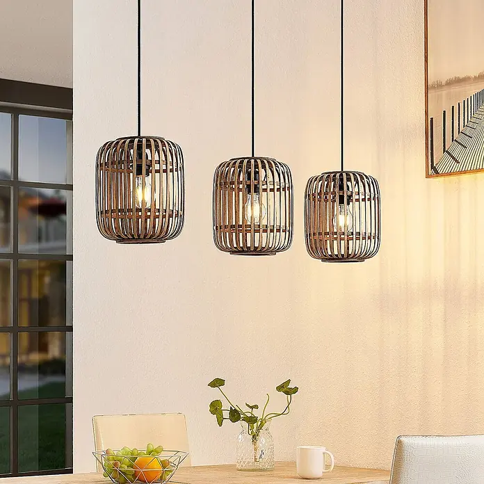 Lindby Pendelleuchte Canyana - L x B x H: 105 x 21,5 x 25 cm, Holz hell, natur, E27 - Drei Pendelleuchten mit natürlichen Bambuskäfigen und dunklen Metallakzenten, die über einem Esstisch hängen.