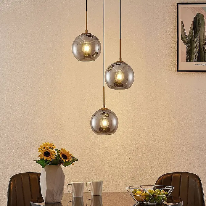 Lindby Pendelleuchte Jurian - L x B x H: 32 x 32 x 17,5 cm, alu / grau / zink, bronze, rauchgrau, E27 - Drei rauchgraue Glaspendelleuchten mit Messingdetails hängen in unterschiedlichen Höhen über einem Esstisch und beleuchten ihn.