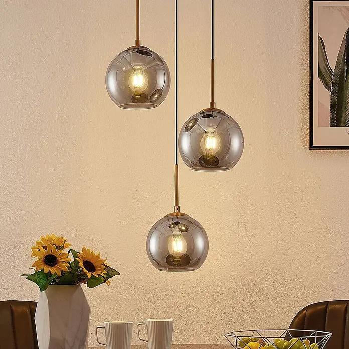 Lindby Pendelleuchte Jurian - L x B x H: 32 x 32 x 17,5 cm, alu / grau / zink, bronze, rauchgrau, E27 - Drei Pendelleuchten mit kugelförmigen Rauchglasschirmen und Messingbeschlägen hängen in unterschiedlichen Höhen über einem Tisch.