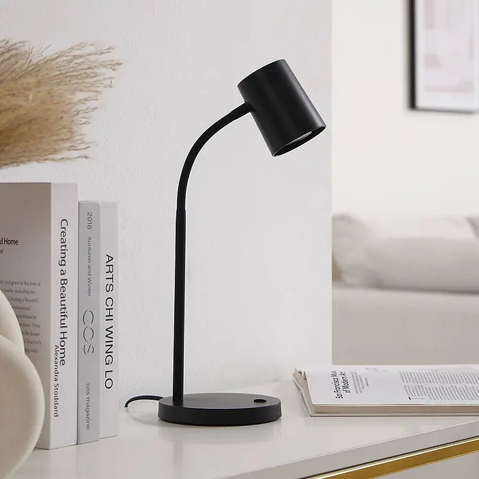 Lindby Tischleuchte Radka - L x B x H: 14,2 x 14,2 x 40,5 cm, schwarz, schwarz, GU10 - Eine schwarze, flexible Schreibtischlampe mit zylindrischem Kopf steht auf einem Tisch neben mehreren Büchern.