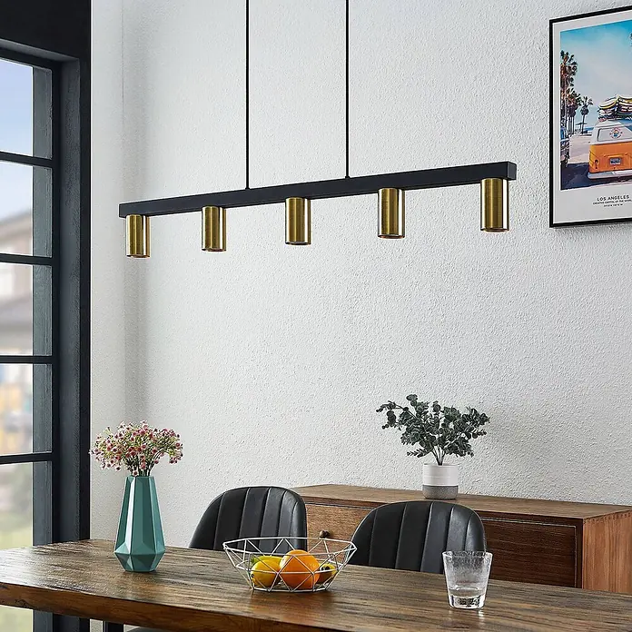 Lindby Pendelleuchte Baliko - messing, Schwarz, GU10 - Eine schwarze lineare Pendelleuchte mit fünf zylindrischen Spotlights aus Messing hängt über einem Esstisch aus Holz.