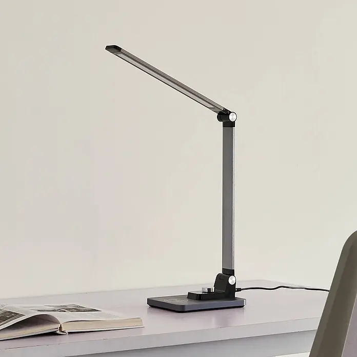 Lindby Tischleuchte Valtaris - L x B x H: 35,6 x 13,7 x 43,1 cm, alu / grau / zink, alu / grau / zink - Eine elegante, dunkelgraue LED-Schreibtischlampe mit verstellbarer Lichtleiste und rechteckigem Sockel steht auf einem Schreibtisch neben einem offenen Buch.