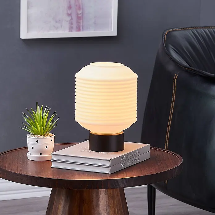 Lindby Tischleuchte Cagla - L x B x H: 18 x 18 x 25 cm, schwarz, weiß / opal, E27 - Eine Tischlampe mit geripptem weißem Glasschirm und dunklem Fuß steht auf einem Holztisch neben einer Topfpflanze und Büchern.
