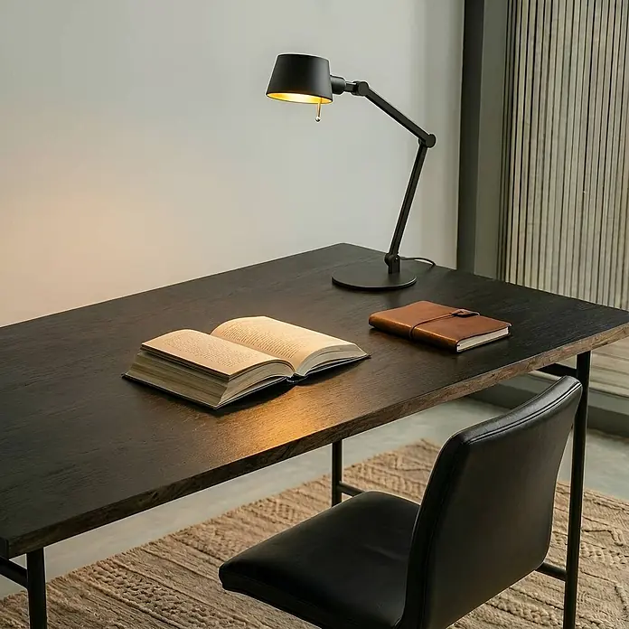 Lucande Tischleuchte Silka - schwarz, schwarz, E27 - Schwarze Metall-Schreibtischlampe mit verstellbarem Arm und goldener Innenseite, beleuchtet einen Holztisch mit Buch und Notizbuch.