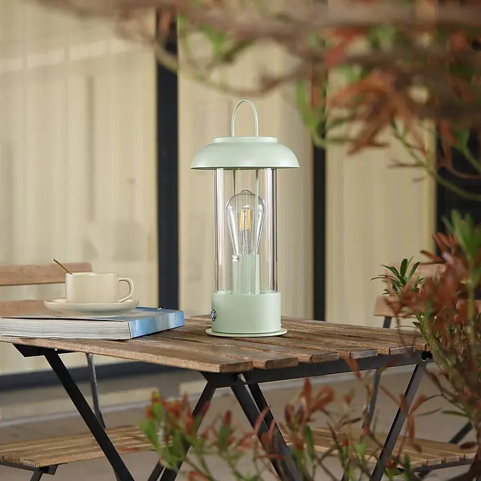 Lindby Tischleuchte Yvette - L x B x H: 17 x 17 x 36,4 cm, grün, klar / transparent, E27 - Mintgrüne tragbare Laterne mit Edison-LED-Birne auf einem Holztisch im Freien neben einer Kaffeetasse und einem Buch.