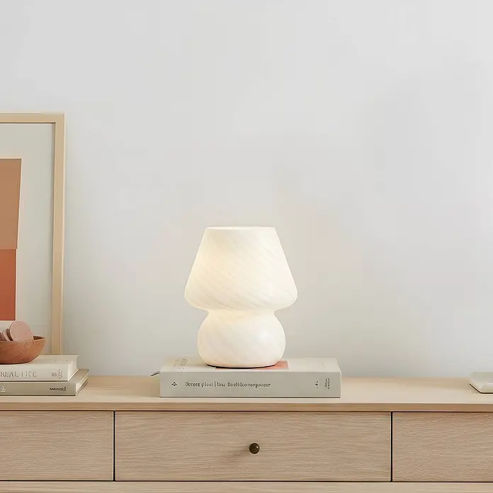 Lindby Tischleuchte Andriana - weiß / opal, weiß / opal, E14 - Weiße, spiralförmige Glas-Pilztischlampe, die einen Raum beleuchtet, steht auf einem hellen Holzschrank neben Büchern.