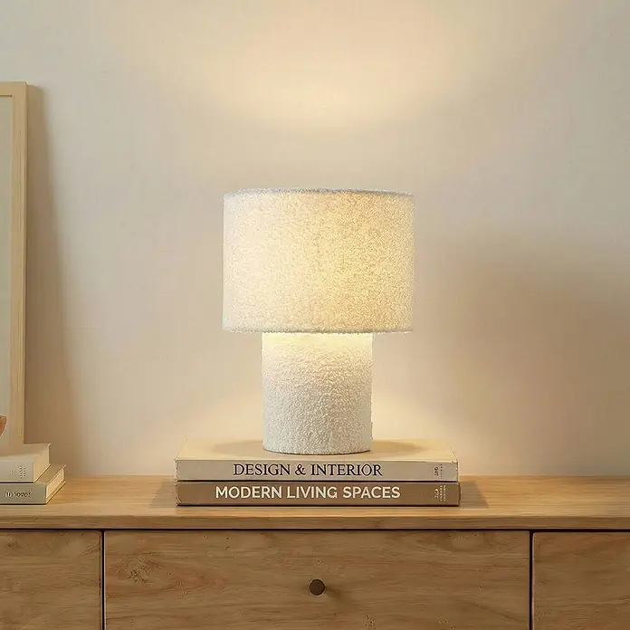 Lindby Tischleuchte Jone - L x B x H: 23 x 23 x 30 cm, weiß / opal, weiß / opal, E27 - Eine weiße, strukturierte Tischlampe mit zylindrischem Fuß und Schirm steht auf einem Holzschrank und strahlt warmes Licht aus.