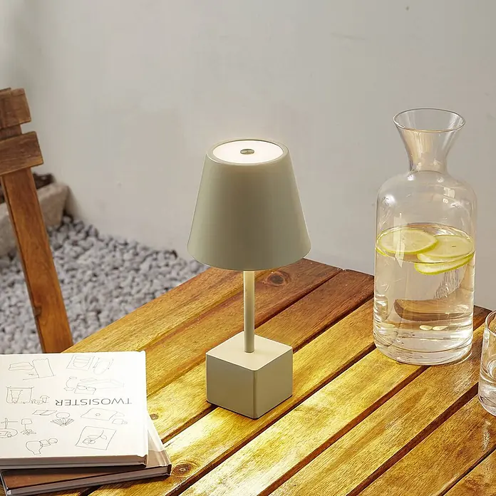 Lindby Tischleuchte Janea - grün, grün - Eine blassgrüne tragbare Tischlampe auf einem Holztisch im Freien, neben einer Karaffe Zitronenwasser und einem Buch.