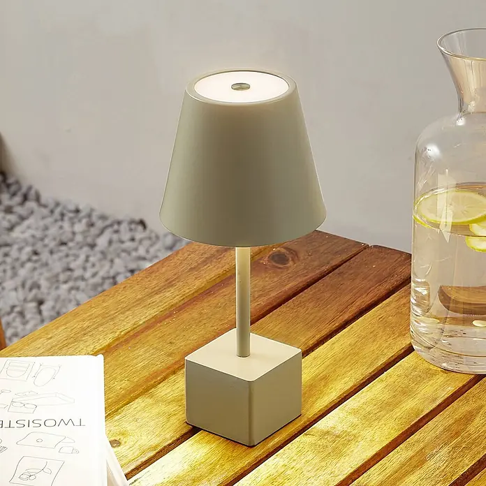 Lindby Tischleuchte Janea - grün, grün - Eine moderne, hellgrüne Tischlampe mit konischem Schirm und quadratischem Fuß steht auf einem Holztisch und spendet Umgebungslicht.