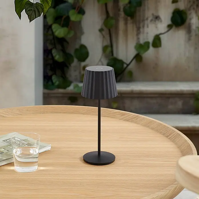 Lindby Tischleuchte Esali - L x B x H: 11 x 11 x 28,5 cm, schwarz, schwarz - Schwarze Tischlampe mit geriffeltem Schirm auf hellem Holztisch im Freien, neben Wasserglas und Magazin.