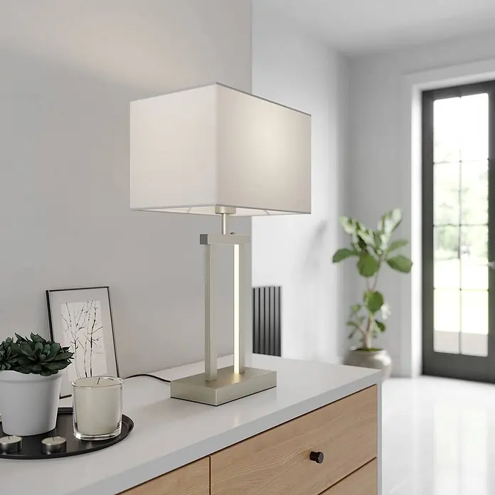 Lindby Tischleuchte Sigurd - alu / grau / zink, weiß / opal, E27 - Moderne Tischlampe mit rechteckigem weißen Schirm und gebürstetem Metallgestell mit integriertem vertikalen LED-Lichtstreifen auf einer Theke.