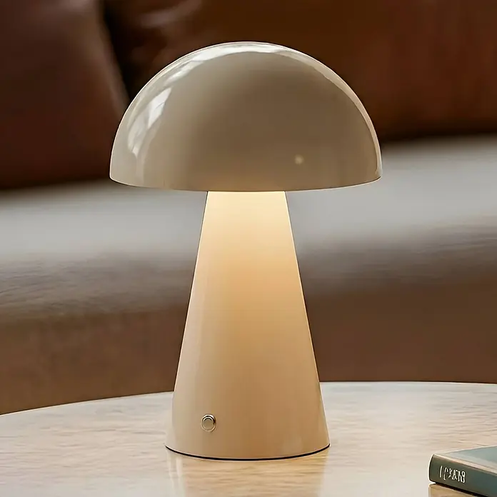 Lindby Tischleuchte Nevijo - L x B x H: 20 x 20 x 30 cm, creme / amber, creme / amber - Pilzförmige, hellbeige Tischleuchte mit glänzender Kappe und sanftem, warmem Licht auf einem hellen Tisch.