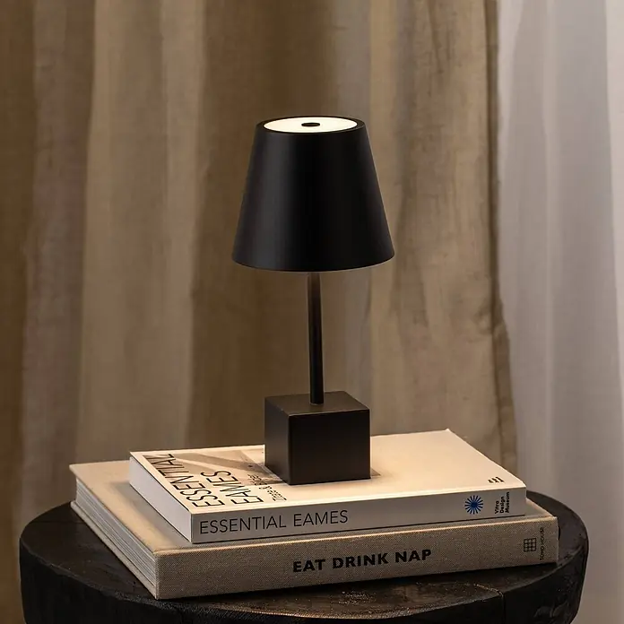 Lindby Tischleuchte Janea - L x B x H: 11,4 x 11,4 x 25 cm, schwarz, schwarz - Schwarze Metall-Tischleuchte mit konischem Schirm und quadratischem Sockel, beleuchtet Bücher auf einem dunklen Holzhocker.