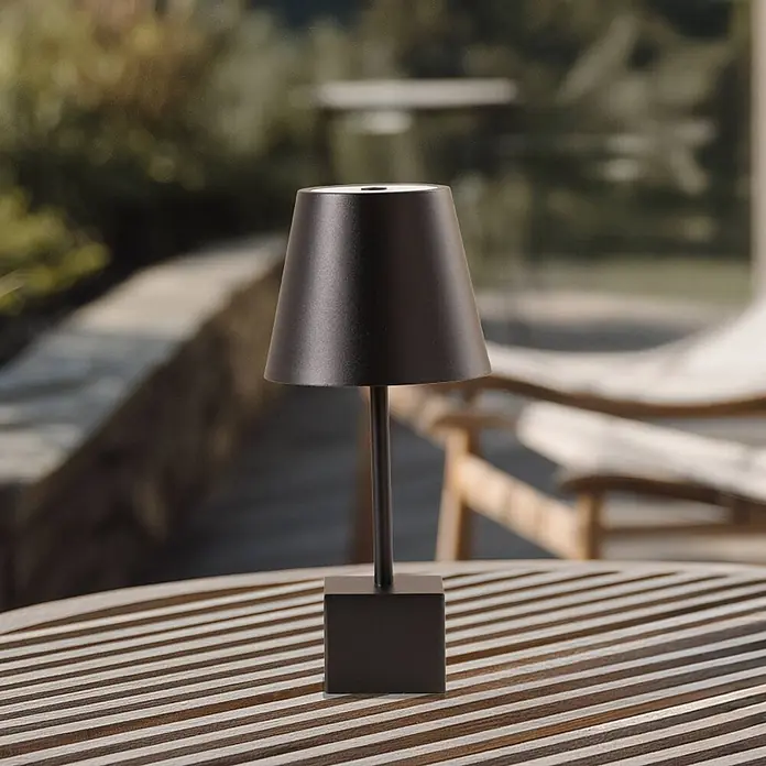 Lindby Tischleuchte Janea - L x B x H: 11,4 x 11,4 x 25 cm, schwarz, schwarz - Eine schwarze tragbare Tischlampe mit konischem Schirm und quadratischem Fuß steht auf einem Lattenholztisch im Freien.
