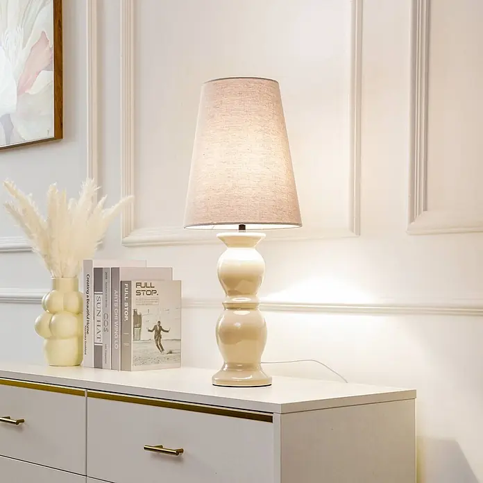 Lucande Tischleuchte Lyndorin - creme / amber, creme / amber, E27 - Cremefarbene Keramiktischlampe mit beigem Stoffschirm spendet gemütliches Licht auf einer Kommode.