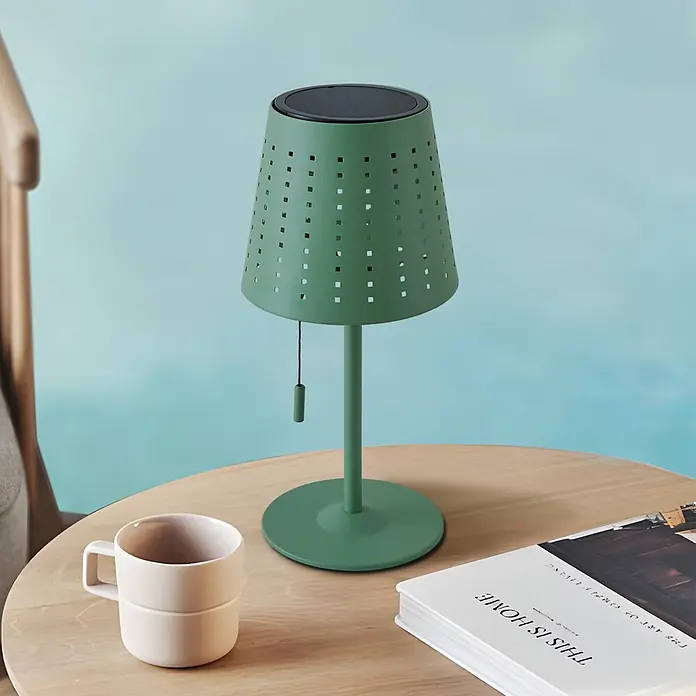 Lindby Tischleuchte Hilario - grün, grün - Grüne perforierte Solar-Tischleuchte mit schwarzer Oberseite auf einem Holztisch neben einer beigen Tasse und einem Buch.