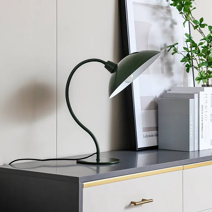 Lucande Tischleuchte Arvadon - grün, grün, E14 - Dunkelgrüne, geschwungene Metall-Schreibtischlampe mit weißem Schirm auf grauer Oberfläche neben Büchern und Bild.