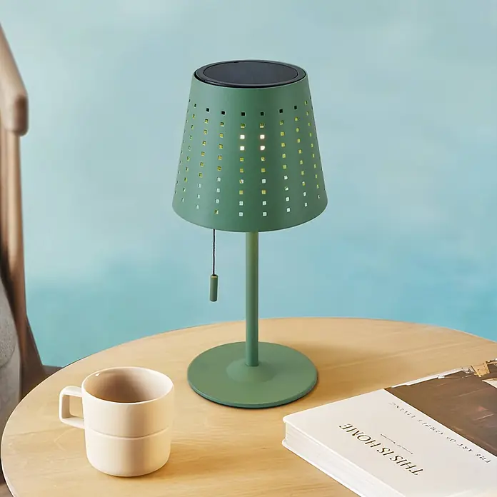 Lindby Tischleuchte Hilario - grün, grün - Grüne Solar-Tischlampe mit perforiertem Schirm, beleuchtet einen Holztisch neben einer beigefarbenen Tasse und einem Buch. Mit Zugschnur.