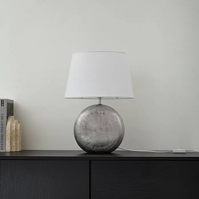 Lindby Tischleuchte Florentino - L x B x H: 35 x 23,5 x 48,5 cm, alu / grau / zink, weiß / opal, E27 - Eine Tischleuchte mit weißem Schirm und strukturiertem, kugelförmigem silbergrauem Fuß auf einem dunklen Holzschrank.