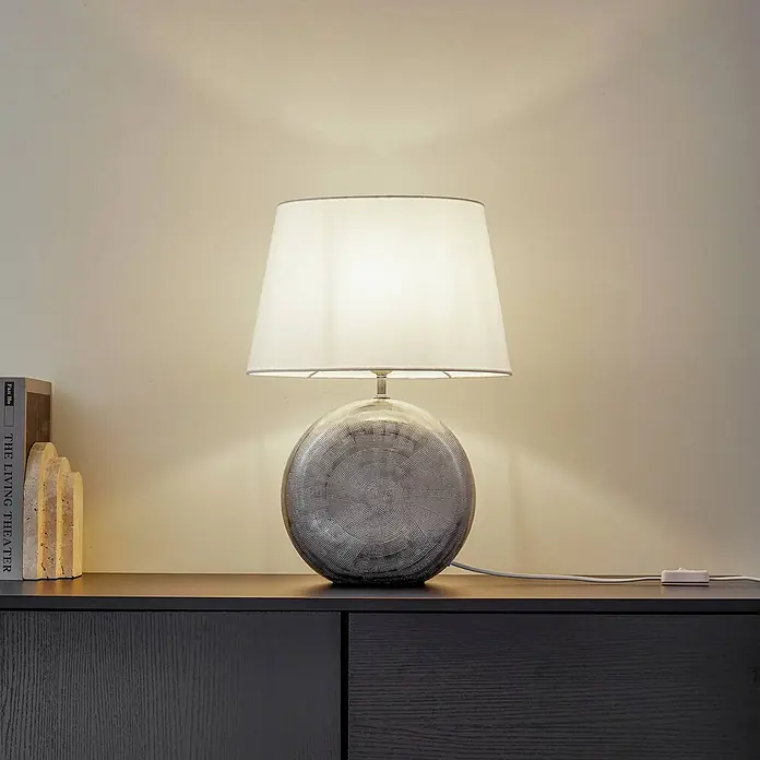 Lindby Tischleuchte Florentino - L x B x H: 35 x 23,5 x 48,5 cm, alu / grau / zink, weiß / opal, E27 - Tischlampe mit rundem, texturiertem grauem Sockel und weißem Stoffschirm, beleuchtet eine Wand.
