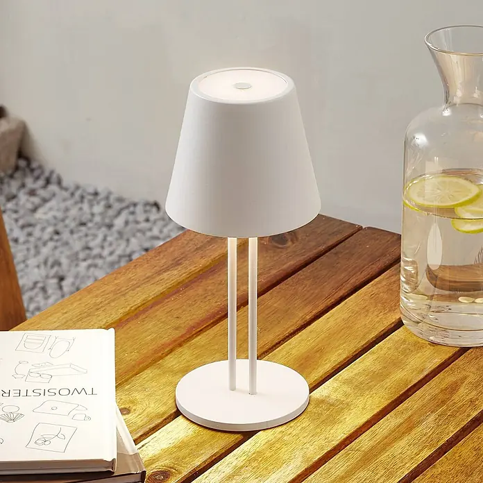 Lindby Tischleuchte Janea - weiß / opal, weiß / opal - Weiße tragbare Tischlampe mit kegelförmigem Schirm und zwei dünnen Stützen auf einem Holztisch.