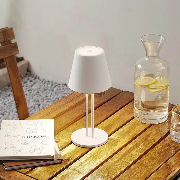 Lindby Tischleuchte Janea - weiß / opal, weiß / opal - Eine weiße tragbare Tischlampe mit konischem Schirm und schlankem Fuß steht auf einem Holztisch neben einer Wasserkaraffe und Büchern.