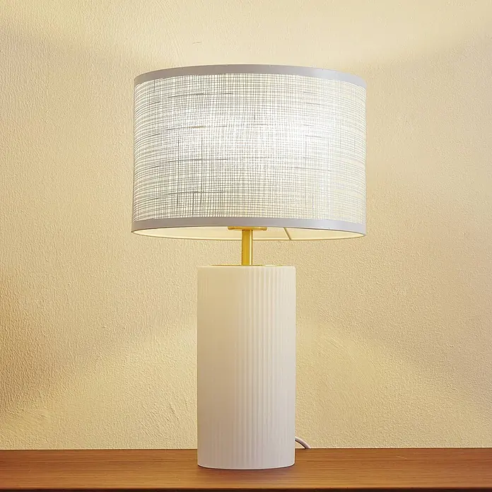 Lindby Tischleuchte Sorscha - L x B x H: 26 x 26 x 43 cm, weiß / opal, weiß / opal, E27 - Beleuchtete Tischlampe mit weißem geriffeltem zylindrischem Fuß, goldenem Stiel und grauem strukturiertem Trommelschirm auf einer Holzoberfläche.