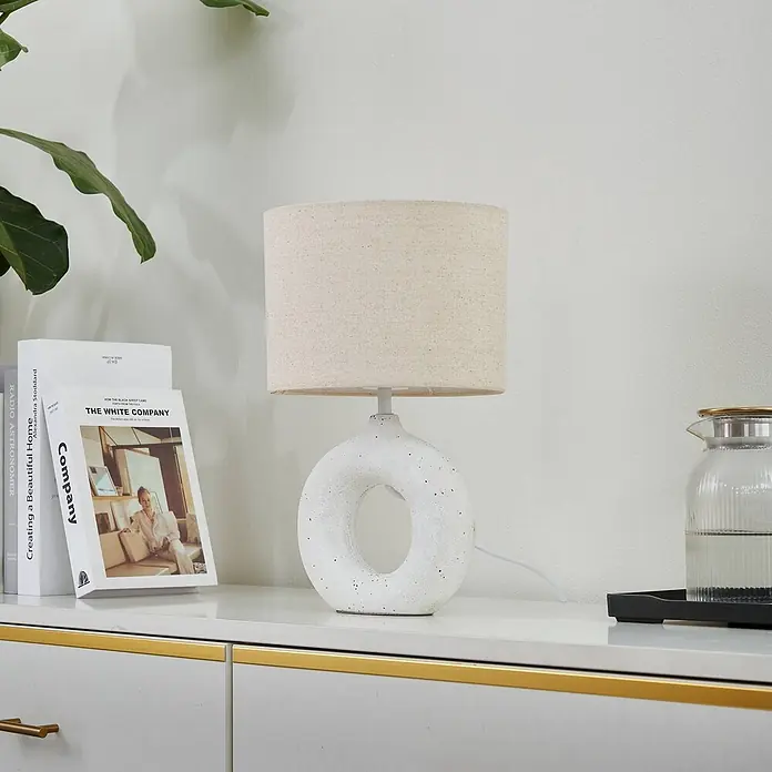 Lindby Tischleuchte Kody - weiß / opal, creme / amber, E27 - Weiße Keramik-Tischlampe in Donut-Form mit Leinen-Schirm, auf einem Schrank.