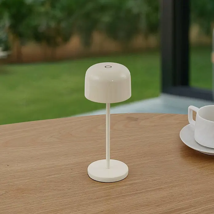 Lucande Tischleuchte Quill - creme / amber, creme / amber - Creme-farbene tragbare Tischlampe auf einem Holztisch, mit einer weißen Teetasse.
