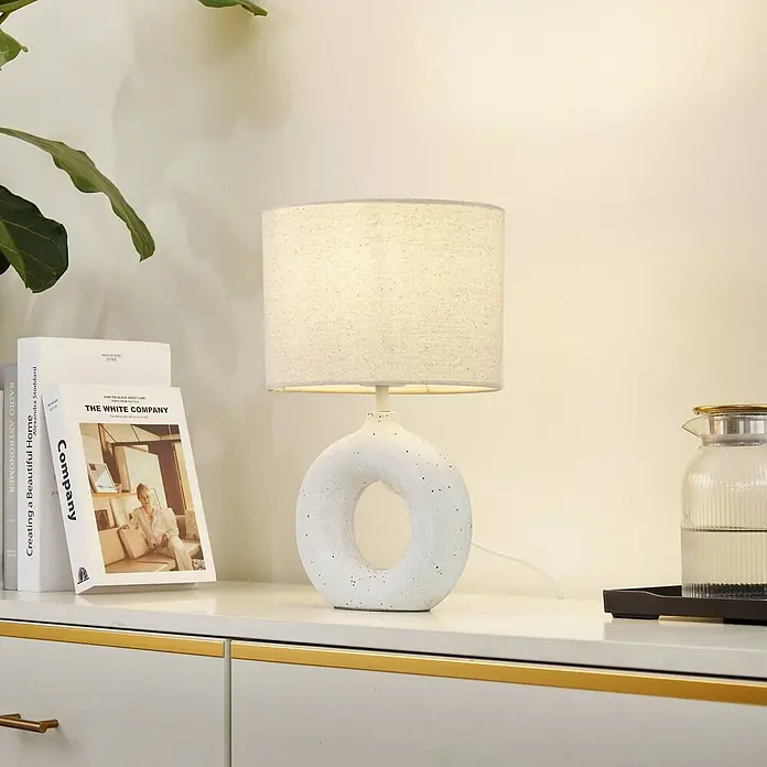 Lindby Tischleuchte Kody - weiß / opal, creme / amber, E27 - Moderne Tischleuchte mit ringförmigem, texturiertem weißem Sockel und hellem Stoffschirm, leuchtet einen weißen Schrank an.