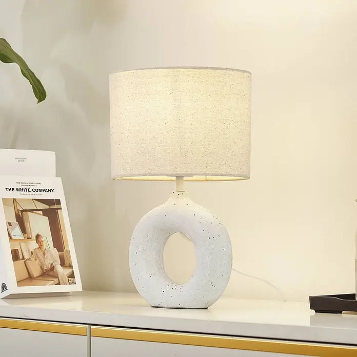 Lindby Tischleuchte Kody - weiß / opal, creme / amber, E27 - Moderne Tischlampe mit Leinen-Schirm und ringförmigem, gesprenkeltem weißem Keramiksockel, beleuchtet auf einer Kommode.