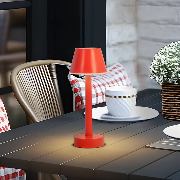 Lindby Tischleuchte Gaja - rot, rot - Eine leuchtend rote Tischlampe beleuchtet einen dunklen Latten-Gartentisch, neben einer weißen Tasse und einer Pflanze platziert.