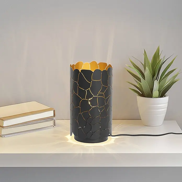 Lindby Tischleuchte Aralena - L x B x H: 15 x 15 x 26,5 cm, schwarz, E27 - Eine schwarz-goldene Tischleuchte mit dekorativem Ausschnittmuster, die Licht auf ein Regal neben Büchern und einer Pflanze wirft.