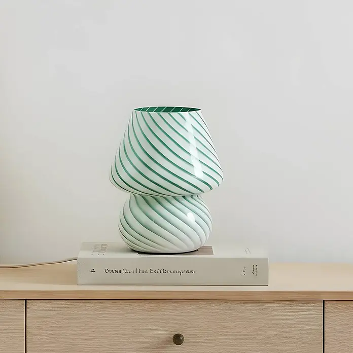 Lindby Tischleuchte Andriana - L x B x H: 15 x 15 x 18,2 cm, grün, grün, E14 - Grün-weiß gestreifte pilzförmige Glas-Tischlampe auf einem Buch auf einem Holztisch.