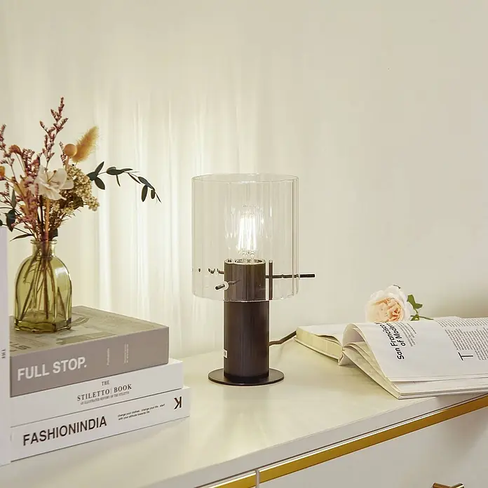 Lucande Tischleuchte Eirian - schwarz, klar / transparent, E27 - Eine Tischlampe mit dunklem Fuß, klarem geriffeltem Glasschirm und sichtbarer Glühbirne auf einem Tisch neben Büchern und Trockenblumen.