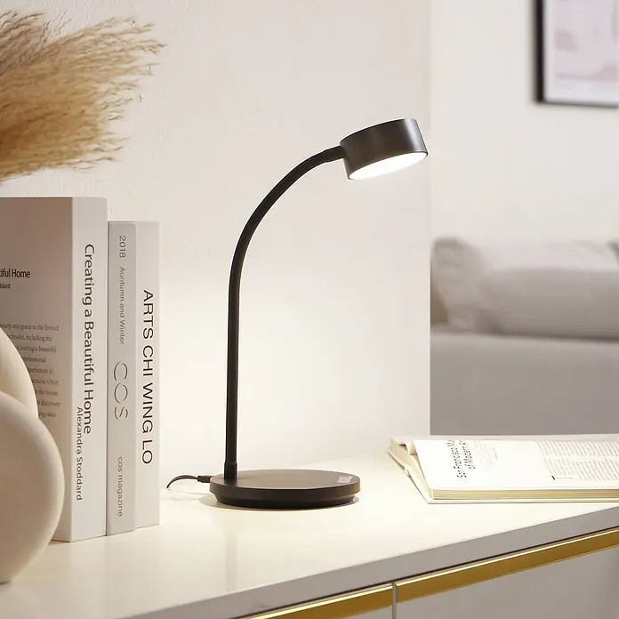 Lindby Tischleuchte Maori - schwarz, schwarz - Eine schwarze Schreibtischlampe mit flexiblem Hals beleuchtet Bücher auf einem weißen Tisch.