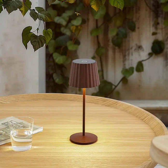 Lindby Tischleuchte Esali - L x B x H: 11 x 11 x 28,5 cm, braun / rost, braun / rost - Braune kabellose Tischlampe mit plissiertem Schirm beleuchtet einen Holztisch im Freien.