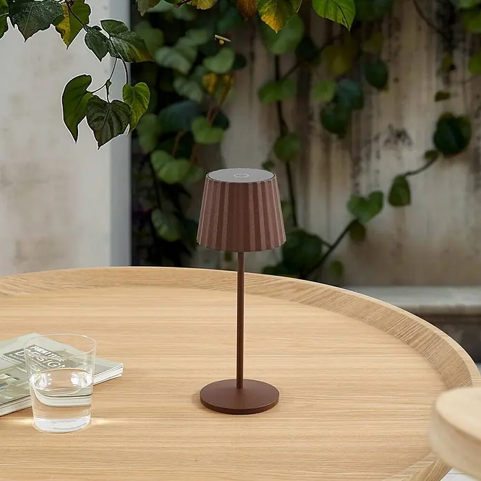Lindby Tischleuchte Esali - L x B x H: 11 x 11 x 28,5 cm, braun / rost, braun / rost - Braune Akkuleuchte mit geriffeltem Schirm auf Holztisch, umgeben von Grünpflanzen.